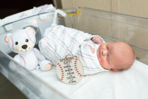 Newborn baby with baseball-themed name sign and teddy bear during Fresh 48 session