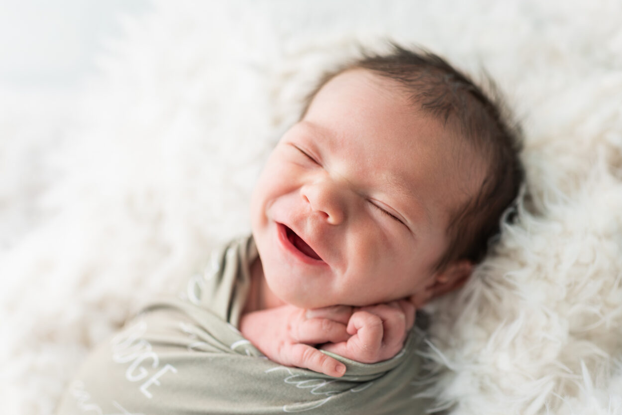 Smiling newborn baby boy wrapped in a sage green swaddle with his name embroidered, on a white fur backdrop