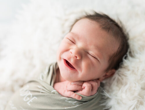 Smiling newborn baby boy wrapped in a sage green swaddle with his name embroidered, on a white fur backdrop