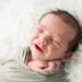 Smiling newborn baby boy wrapped in a sage green swaddle with his name embroidered, on a white fur backdrop