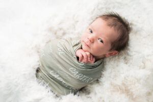 Wide-awake newborn baby boy wrapped in a sage green personalized name swaddle on a white fur backdrop