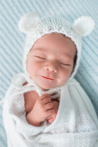 Smiling newborn baby boy wearing a white knit bear hat on a light blue backdrop
