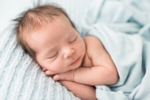 Newborn baby boy sleeping peacefully on a light blue blanket with his hands tucked under his chin