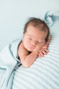 Newborn baby boy resting on a light blue blanket with arms tucked, full head of hair visible