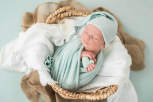 Newborn baby boy swaddled in sage green wrap and hat, sleeping in a woven basket holding a small heart