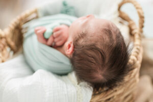 Close-up of sleeping newborn baby's dark hair and sage green swaddle in a woven basket
