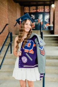 Knoxville senior photographer captures high school senior in navy graduation cap holding a purple and gray varsity letterman jacket on the front steps of her school