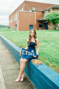 Senior girl in navy dress sitting on a painted wall holding an ETSU pennant and gold pom poms, photographed by a Knoxville senior photographer