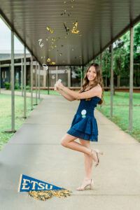 Senior girl popping gold confetti under a school breezeway with an ETSU pennant on the ground, Knoxville senior photographer session