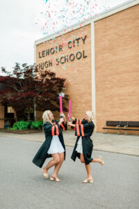 Two graduating seniors celebrating with confetti poppers in caps and gowns outside Lenoir City High School