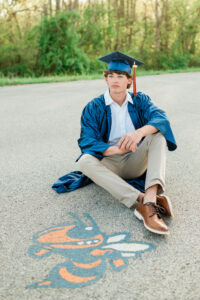 Knoxville senior photographer photo of a high school senior boy sitting on a road in his blue cap and gown beside a hand-painted yellow jacket mascot logo, with a 2024 tassel and green trees in the background.