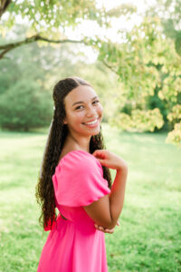 Knoxville senior photographer portrait of a high school senior girl in a bright pink off-shoulder dress smiling over her shoulder under dappled tree light in a green park.