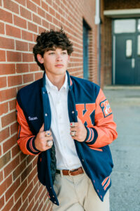 Knoxville senior photographer photo of a high school senior boy leaning against a brick wall wearing a navy and orange varsity letterman jacket embroidered with his name, showing off championship rings.