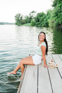 Knoxville senior photographer photo of a high school senior girl in a mint green top and white shorts sitting on a lake dock with her bare feet splashing in the water, sandals beside her.