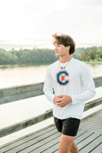 Knoxville senior photographer photo of a high school senior boy in a white long-sleeve athletic shirt and gold chain walking along a lakeside dock at golden hour.