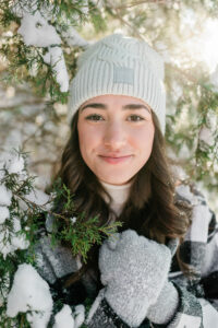 Close-up portrait of senior in white beanie surrounded by snow-covered pine branches