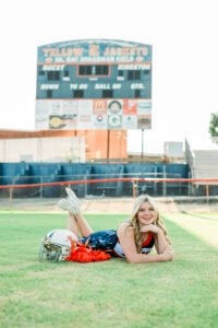 Knoxville senior photographer photo of a high school senior cheerleader lying on the football field in her navy and orange uniform beside a football helmet and pom-poms, with the Yellow Jackets scoreboard in the background.