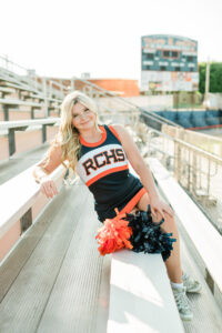 Knoxville senior photographer photo of a high school senior cheerleader in an RCHS navy and orange uniform sitting on stadium bleachers with pom-poms and the football scoreboard blurred behind her.