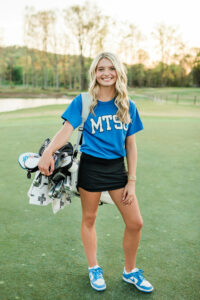 Senior in blue MTSU shirt carrying a golf bag on the golf course at sunset