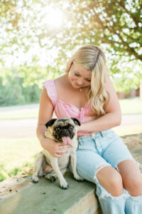 Knoxville senior photographer photo of a high school senior girl in a pink ruffle top and ripped jeans sitting on a stone ledge, laughing while holding her pug under a shady tree.