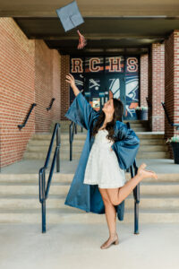 Roane County High School senior girl in a navy cap and gown over a white dress tossing her graduation cap in the air in front of the RCHS entrance doors