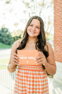 Roane County High School senior girl in an orange gingham dress smiling and holding a "Congrats New Vol" University of Tennessee acceptance card