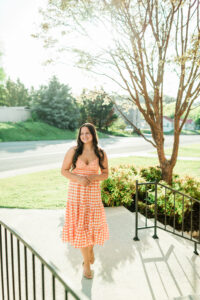 Roane County High School senior girl in an orange and white gingham dress walking along a sidewalk beside a tree and wrought iron railing