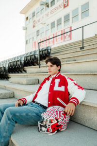 Knoxville area senior in red and white football letterman jacket sitting on stadium steps with helmet, home of the Redskins press box in background