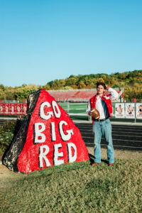 Senior football player holding a football beside a Go Big Red painted spirit rock at the stadium