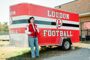 Senior in varsity letterman jacket leaning against the Loudon Football equipment trailer