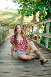 Senior in floral top and denim shorts sitting on a wooden bridge at Southwest Point in Kingston TN wearing cowboy boots