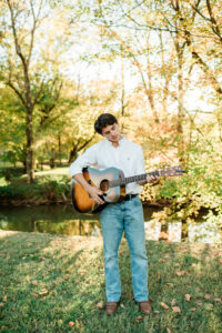 Senior playing acoustic guitar beside a pond surrounded by fall foliage in Loudon TN