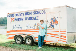 Roane County High School senior boy in a light blue patterned shirt playing a silver trumpet beside the Roane County High School Marching Band equipment trailer in Kingston, Tennessee