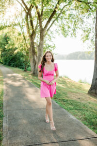 Senior in hot pink dress walking along a lakeside path lined with trees